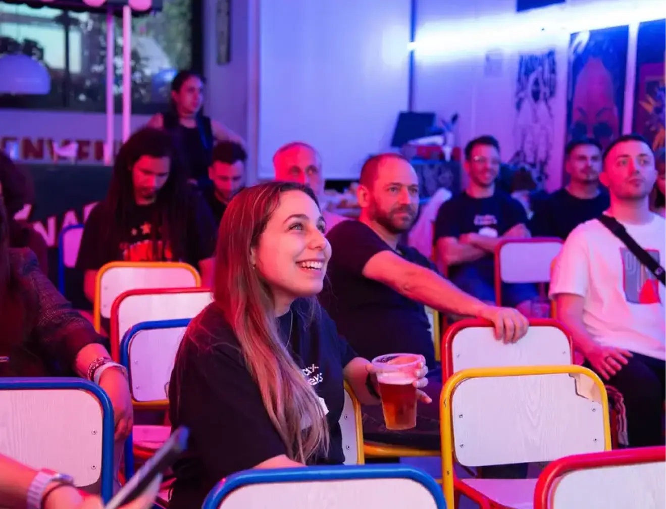 Smiling woman holding a drink and sitting among other attendees at a colorful, neon-lit tech meetup or networking event.