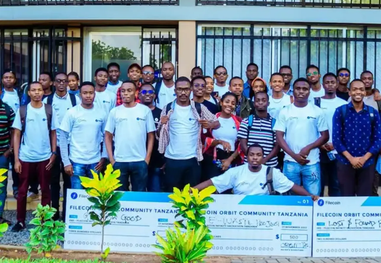 A large group of young people wearing matching white shirts posing for a photo in front of a building during a tech community event, holding a giant presentation check.