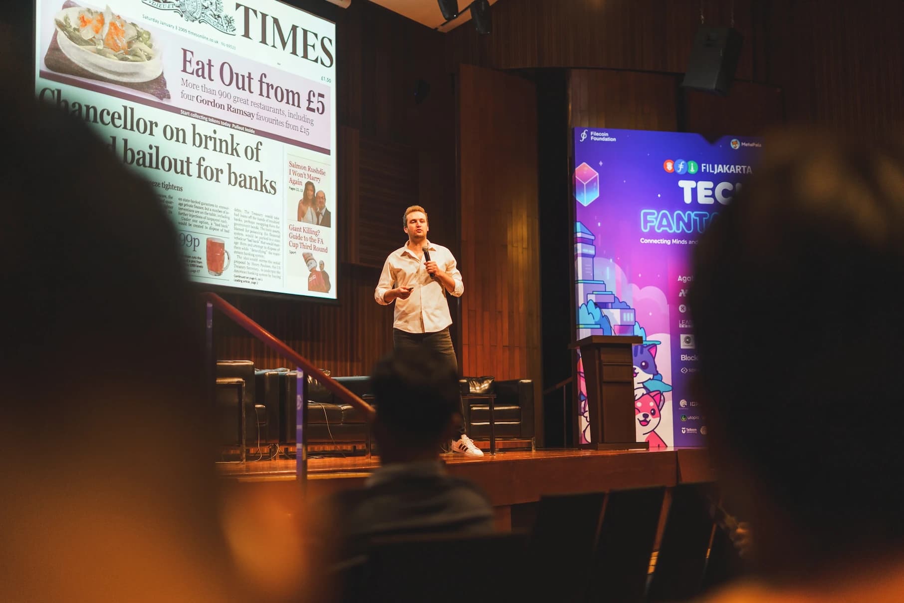 A man giving a keynote presentation on stage at a tech conference, with a projected newspaper slide and a colorful event banner in the background.
